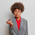 confused puzzled female director, owner of business company, shrugs shoulders with puzzlement, dressed formally, wears round glasses, stands against grey studio background, has no idea what happening