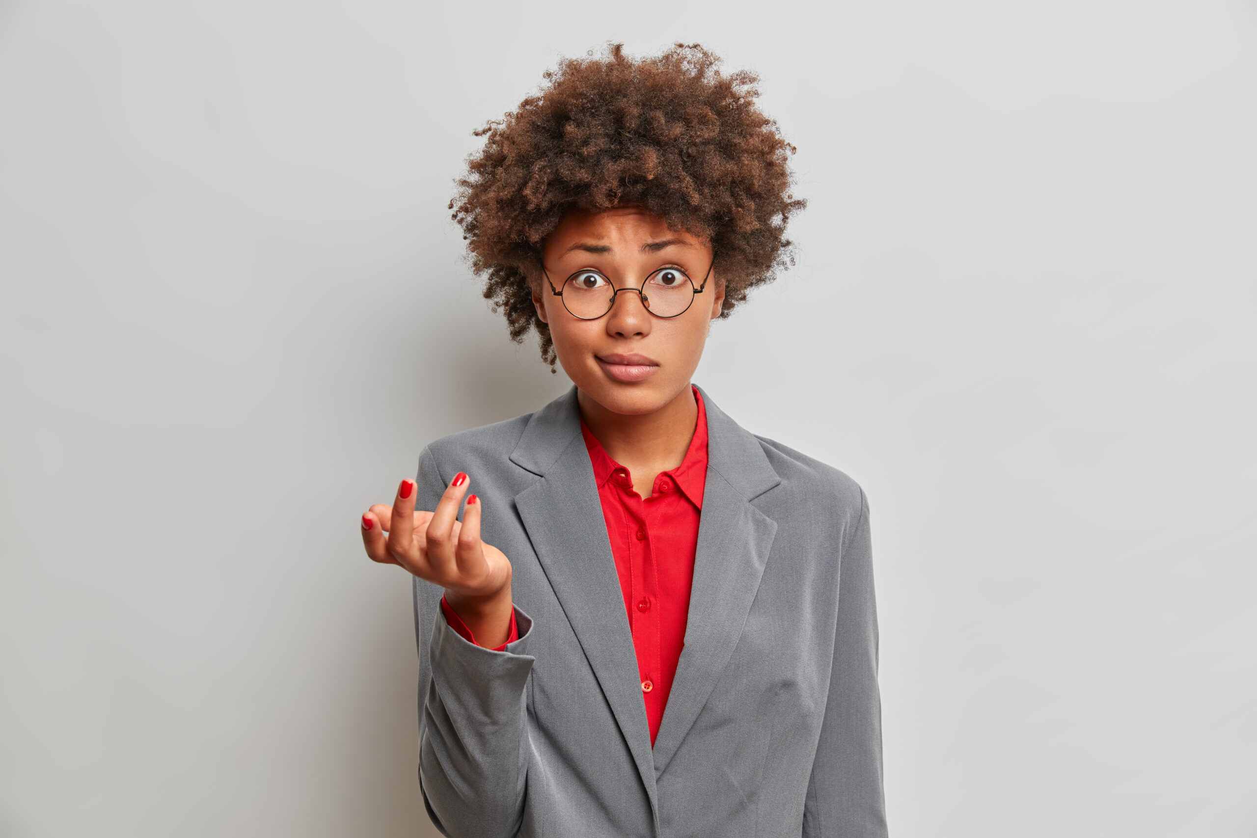 confused puzzled female director, owner of business company, shrugs shoulders with puzzlement, dressed formally, wears round glasses, stands against grey studio background, has no idea what happening